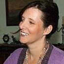 A person smiles while looking to the side, wearing a purple top and beaded necklace, in a room with framed photos and a candlestick.