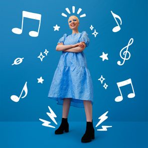 Person smiling confidently, standing with arms crossed, wearing a textured blue dress and black boots, surrounded by white musical notes and stars, against a blue background.