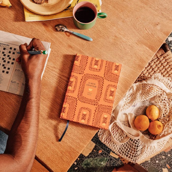 Hand writes crossword on newspaper; notebook and coffee nearby; tote contains peaches on wooden table.
