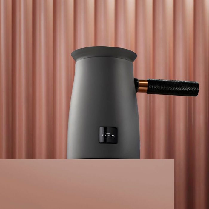 Electric kettle with textured handle sits on table against rippled, pink backdrop. Plaque reads 