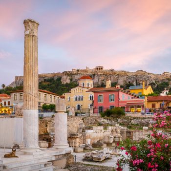 Ancient stone columns stand amidst ruins, with colorful buildings and a hilltop structure in the background, under a vibrant sunset sky.