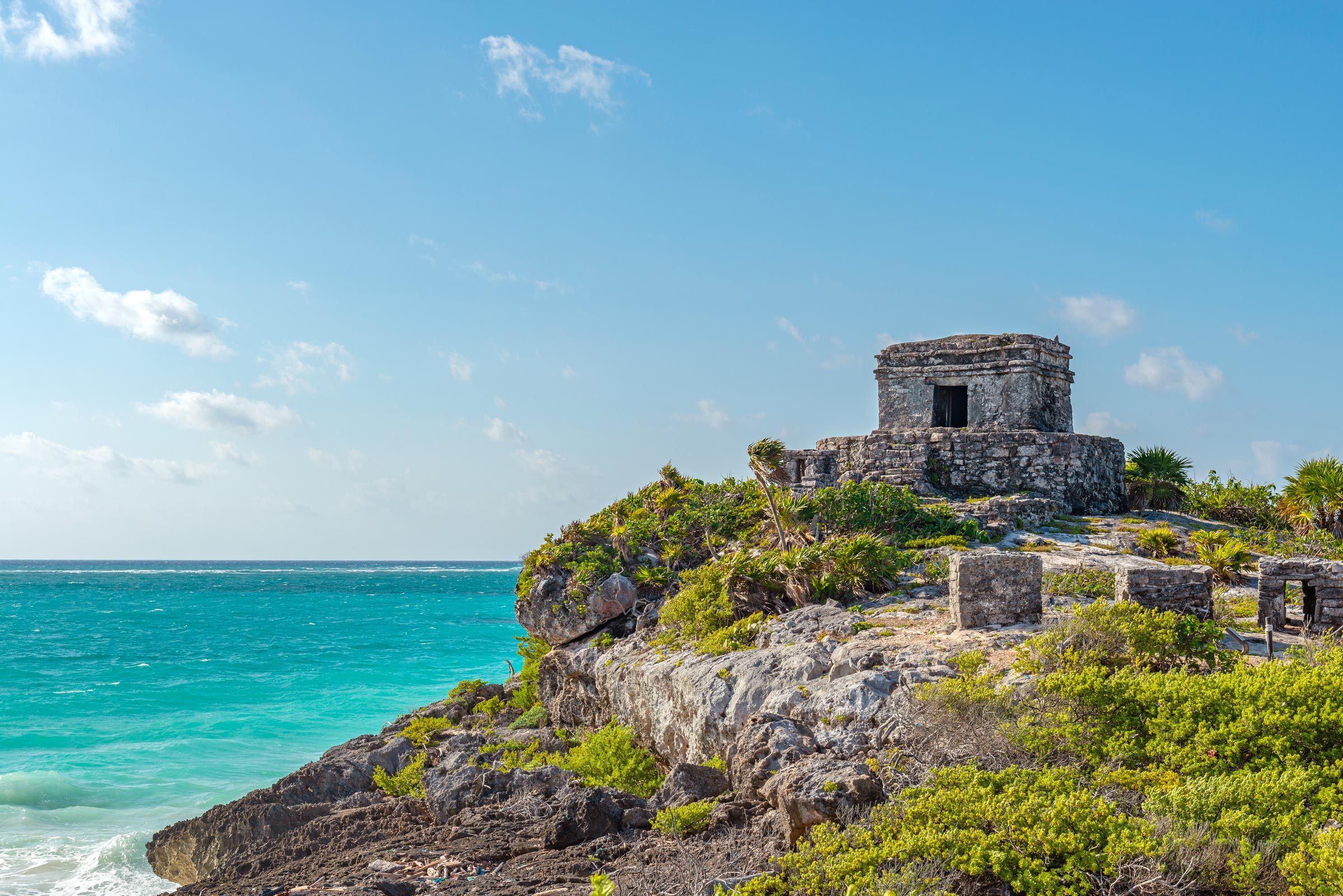 Stone ruins rest on a cliff above turquoise sea, surrounded by lush greenery under a clear blue sky.