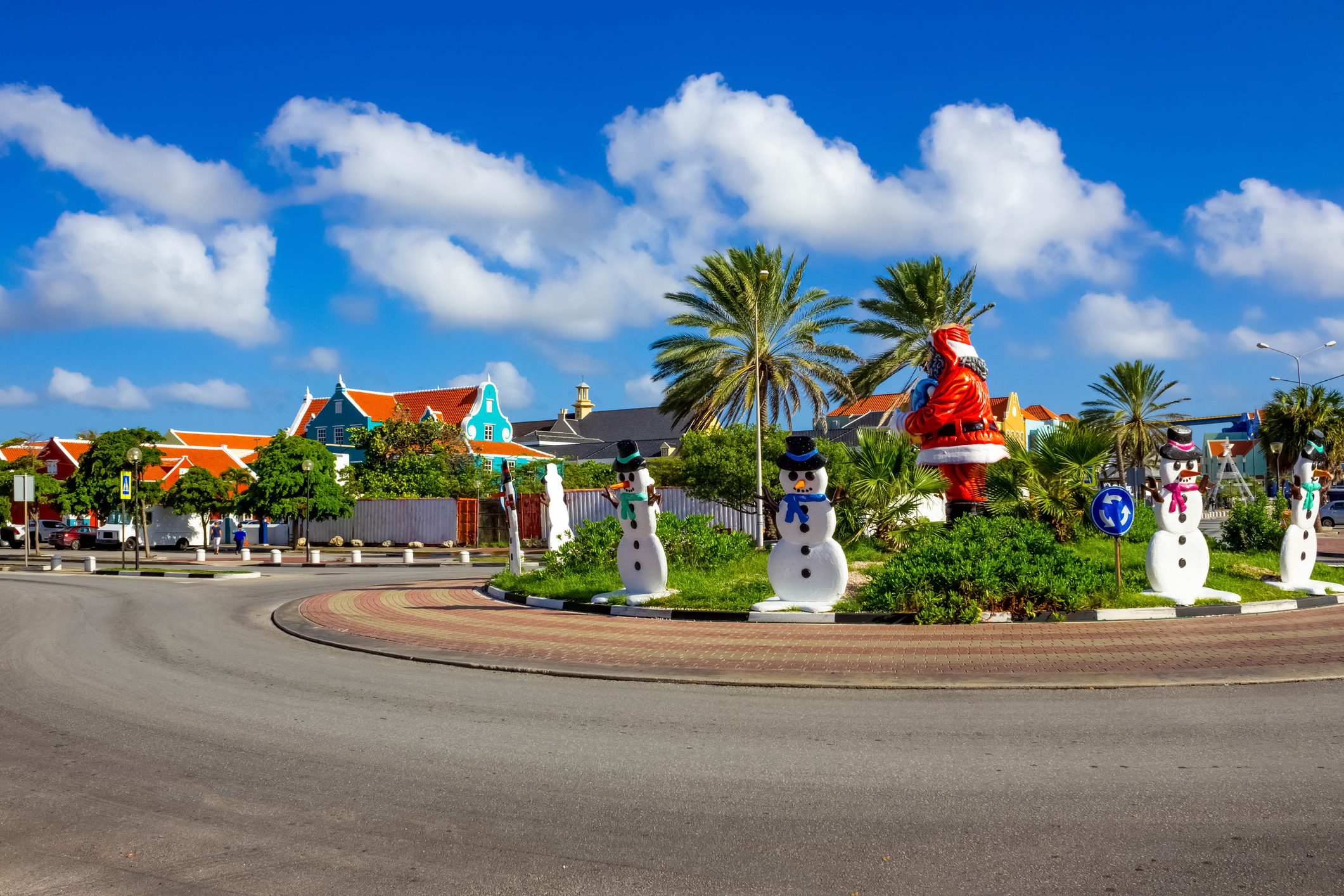 Snowman figures stand in a sunny roundabout, surrounded by palm trees and colorful buildings under a clear blue sky.