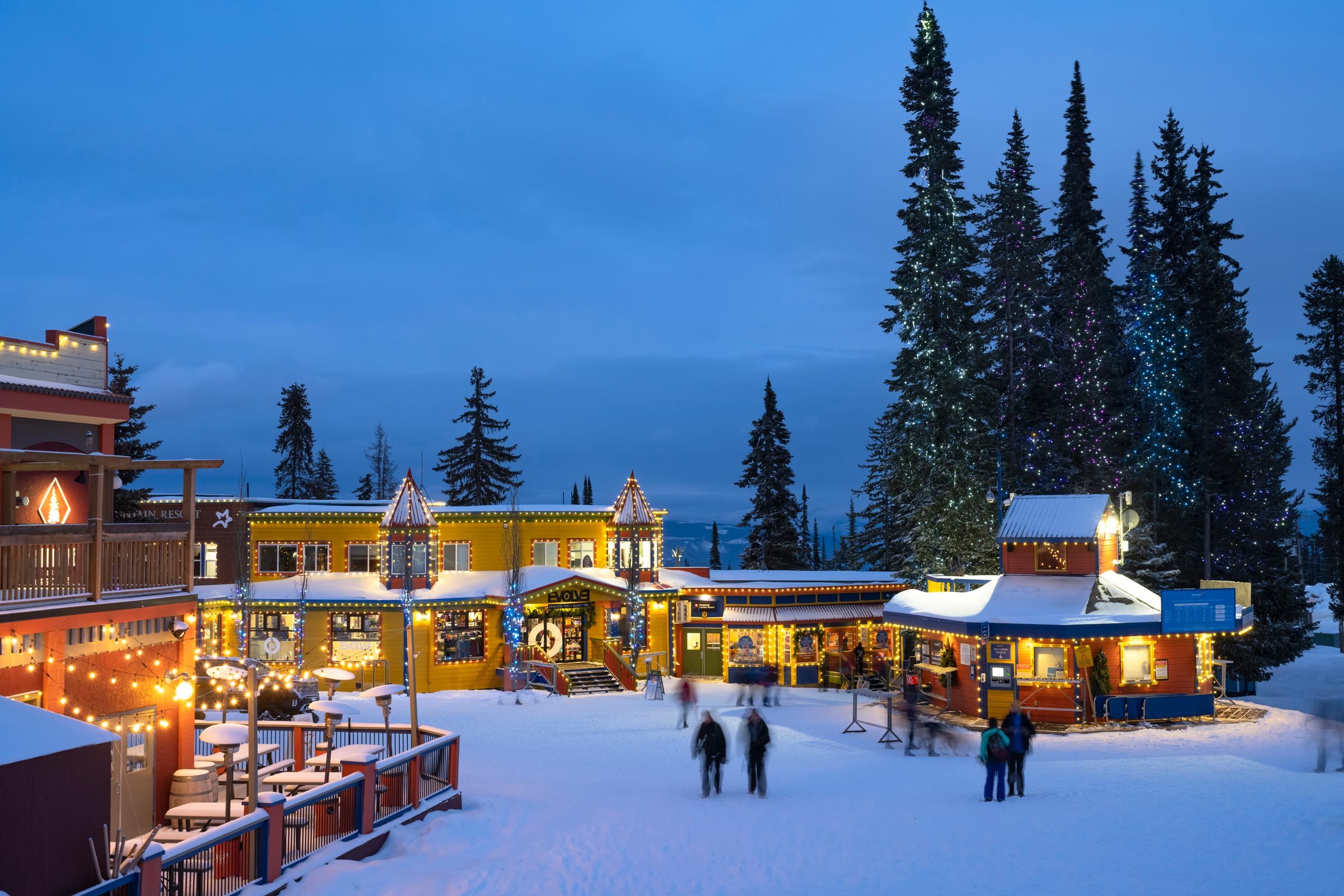 Buildings decorated with festive lights stand in a snowy village, with people walking around; tall trees and a darkening sky provide the backdrop.