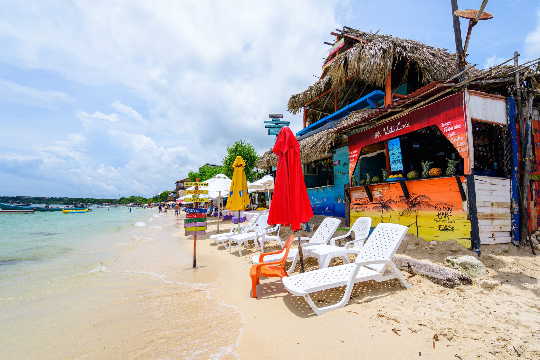Colorful beach bar with thatched roof, chairs, and umbrellas; lively beachfront with boats and people enjoying the water. Sign reads "BAR Vista Linda."
