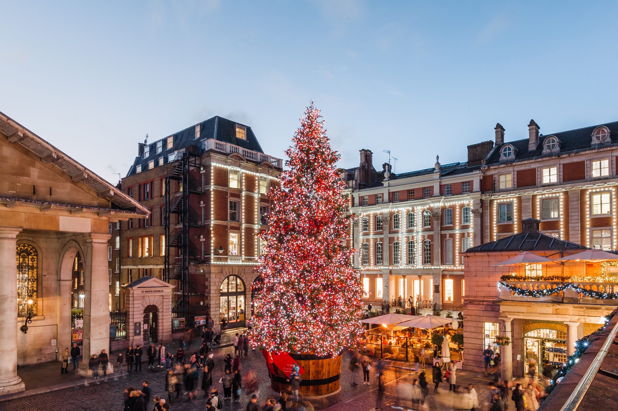Christmas tree illuminated with red and white lights, standing in a busy cobblestone square, surrounded by historic buildings and evening shoppers.