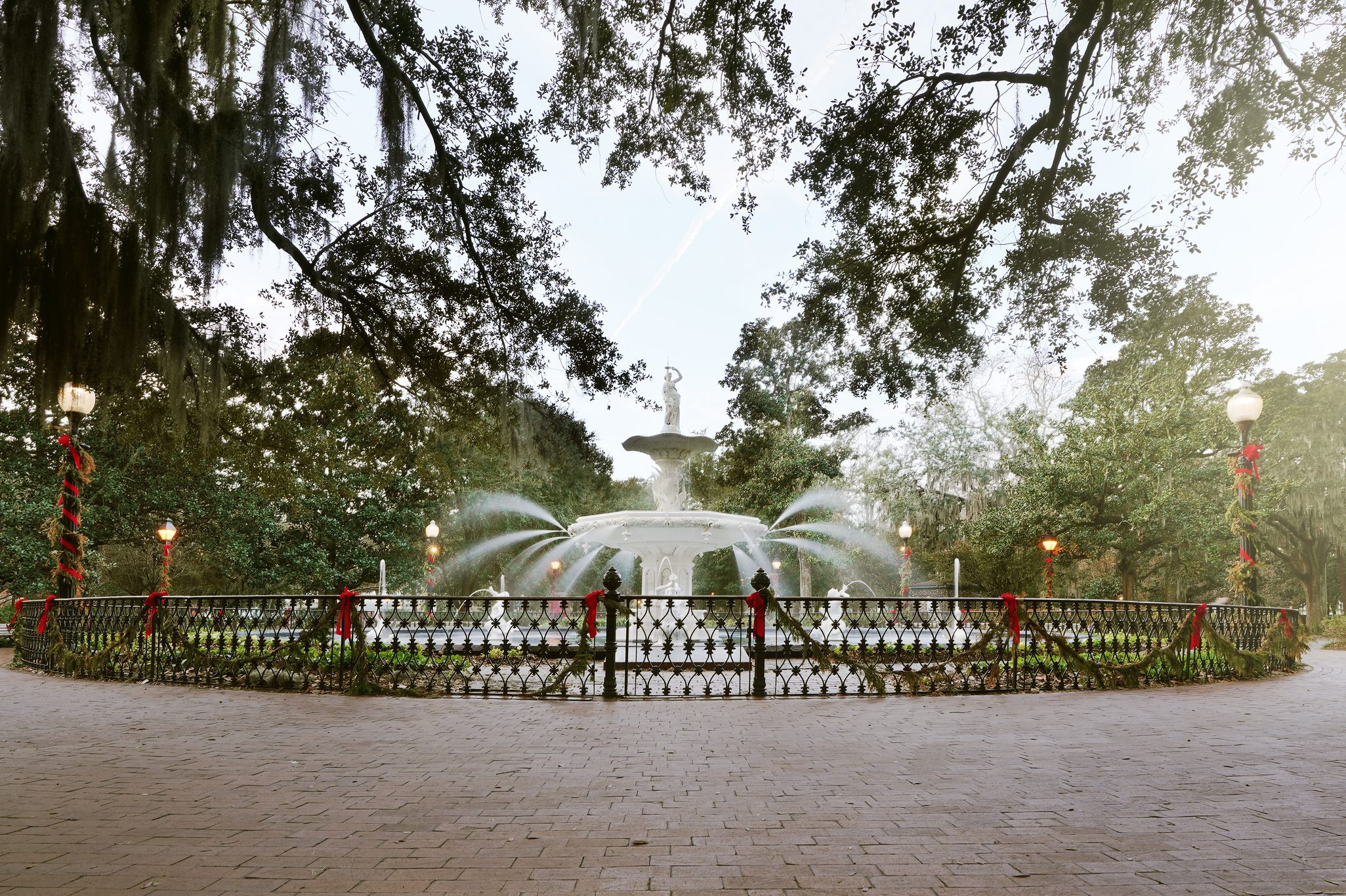 Fountain sprays water in a park surrounded by decorated lampposts and trees, with a paved walkway in the foreground.