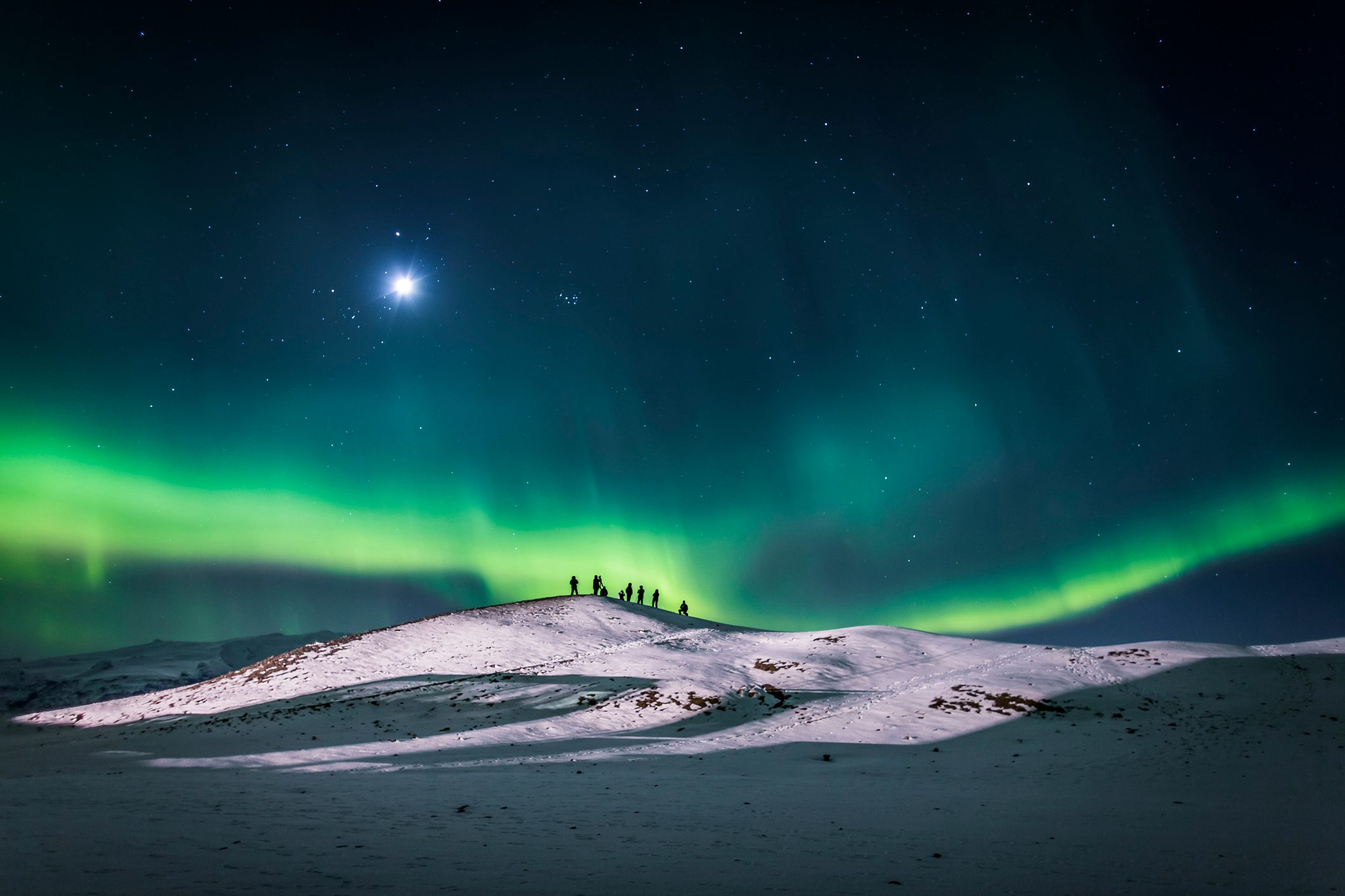 Silhouetted figures stand on a snowy hill, watching the vivid green aurora borealis below a starry night sky.
