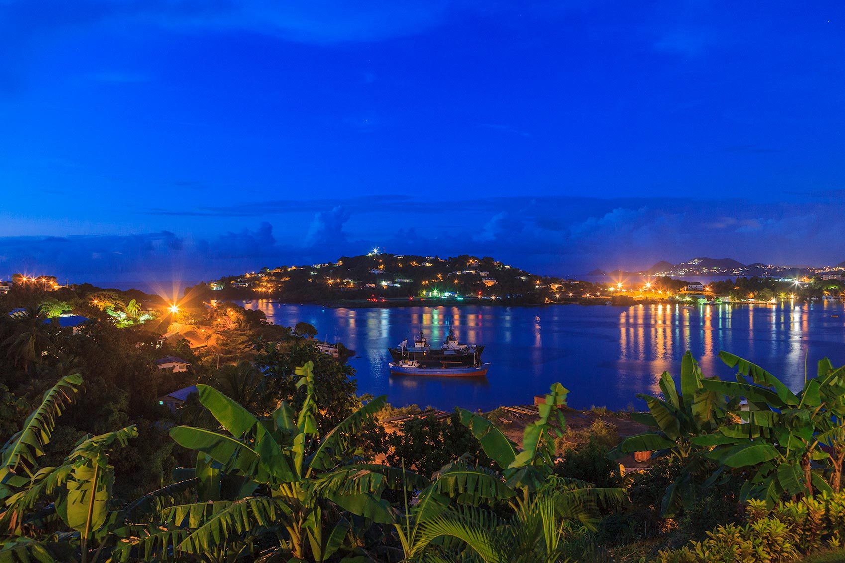 A boat rests on a calm, illuminated bay at night, with verdant foliage and distant glowing lights on surrounding hills.