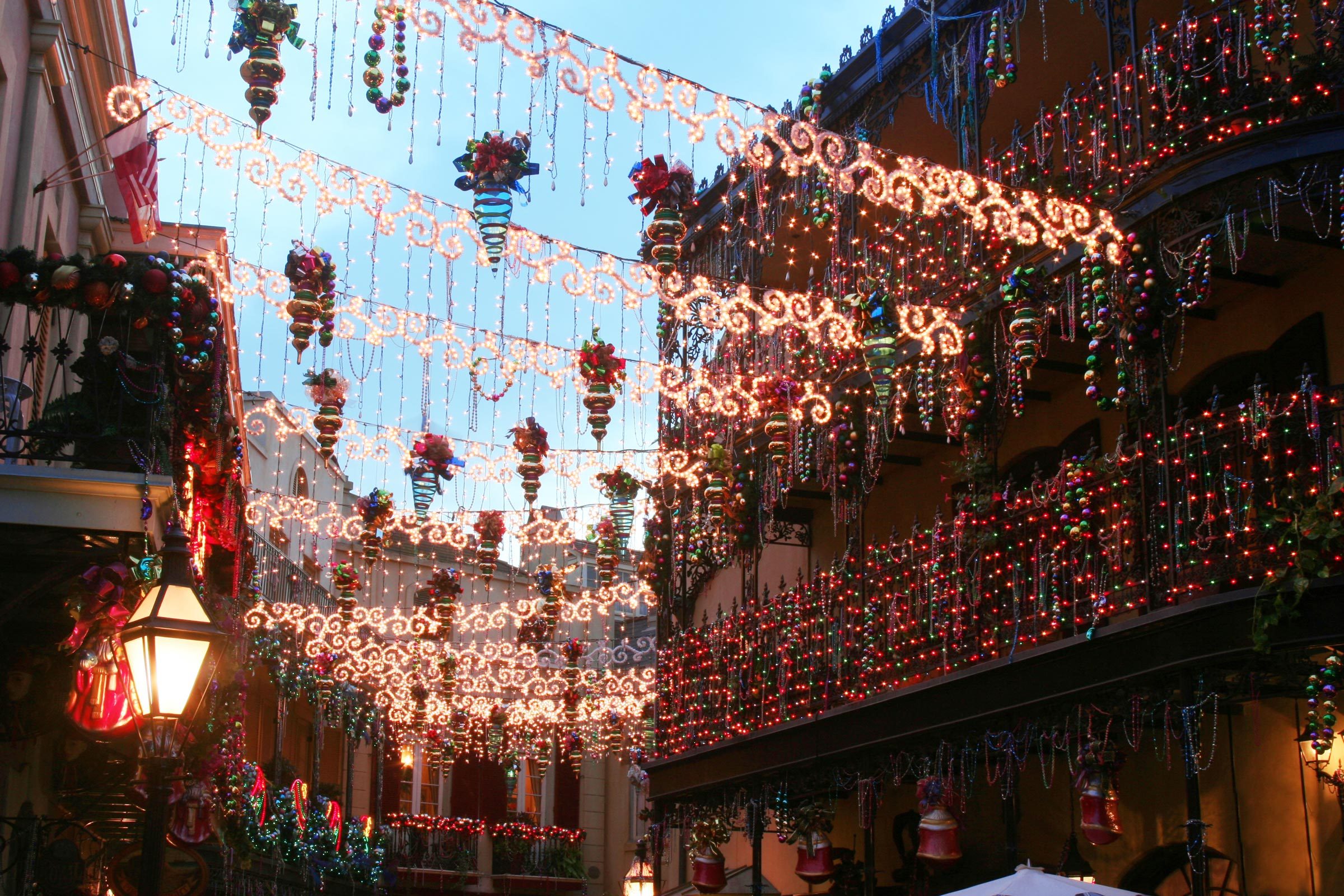 Festive lights adorn a narrow street, hanging in intricate patterns, surrounded by balconies decorated with garlands and colorful ornaments under a clear evening sky.