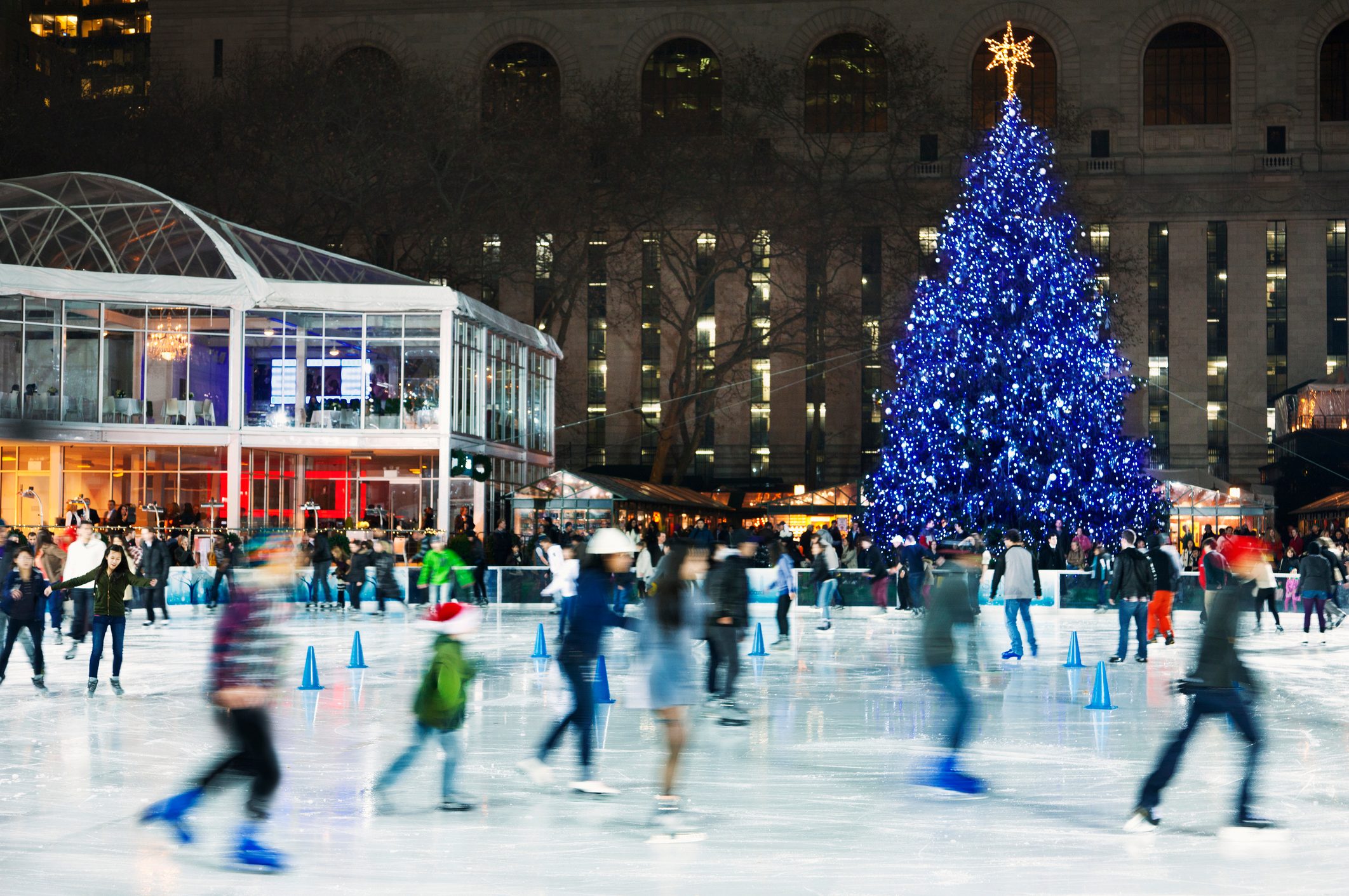 People ice-skating under festive lights near a large, illuminated Christmas tree. A glass pavilion and city buildings surround the busy, lively rink.