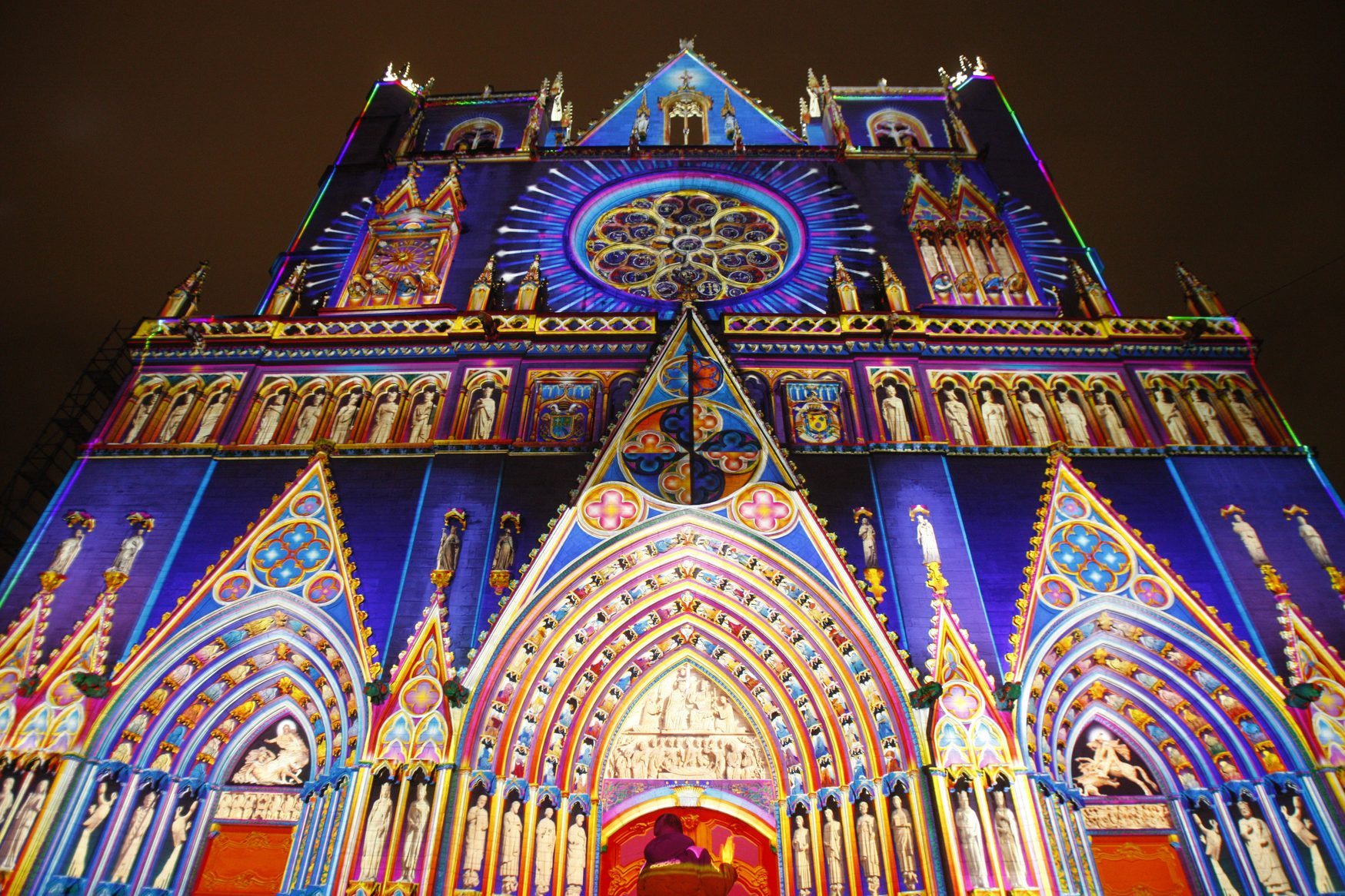 Gothic cathedral facade illuminated, showcasing vibrant colors and intricate patterns at night, highlighting detailed architectural sculptures and elements.