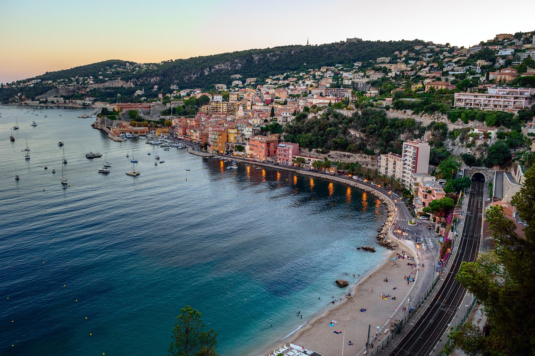 Coastal village sits along a curved shoreline. Boats float in calm bay. Hills with houses rise in the background. Railway tracks run adjacent.