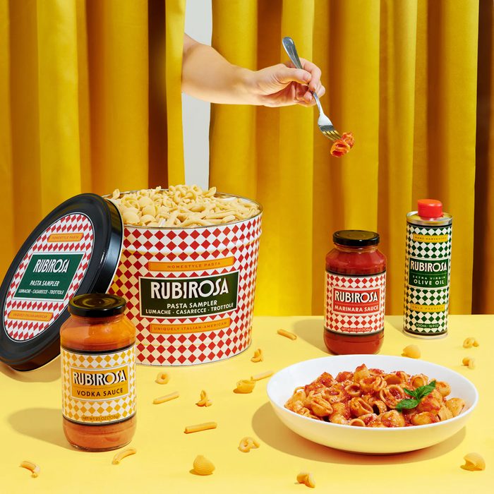 Hand twirls pasta over a bowl on a yellow table. Containers of Rubirosa pasta, sauces, and olive oil are arranged nearby.