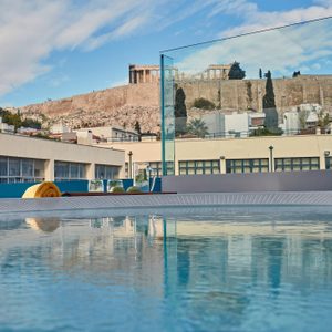 Pool reflects sky, glass pitcher on a table, rooftop setting with Acropolis view in the background.