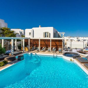 A pool reflecting sunlight lies surrounded by lounge chairs, modern architecture, and clear blue skies, evoking a serene and luxurious urban getaway.