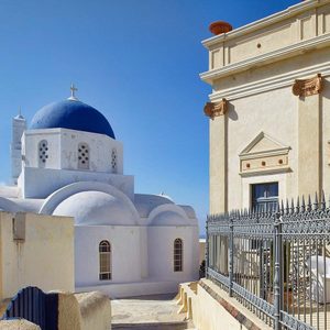 A white building with blue dome stands in sunlight, adjacent to a beige ornate building, surrounded by a clear blue sky.