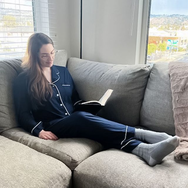 Person reads on a grey couch, wearing navy pajamas and socks, in a bright room with large windows overlooking a scenic view.