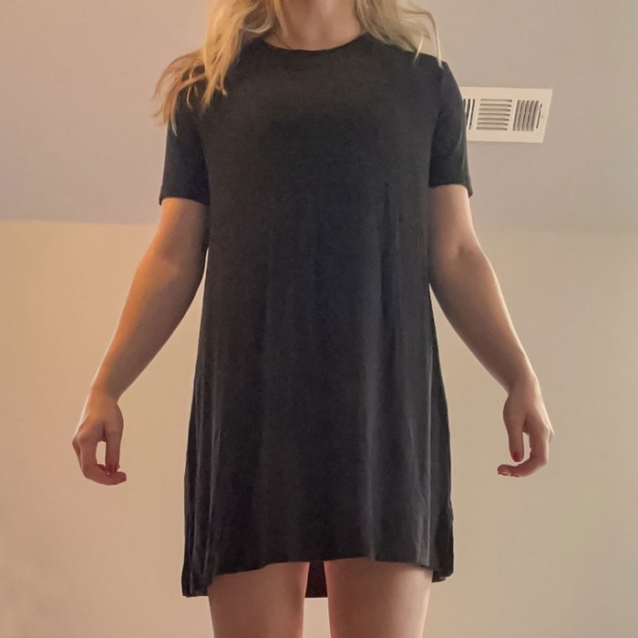 Person wearing a black dress stands with arms relaxed, in a dimly lit room with a visible ceiling vent.