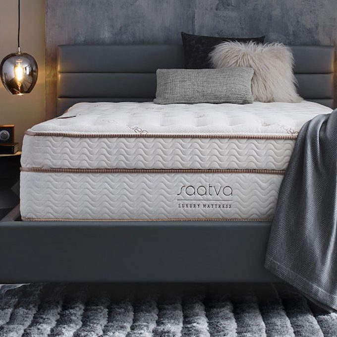 Mattress sits on a sleek dark bed frame, surrounded by cozy pillows and a gray blanket; 