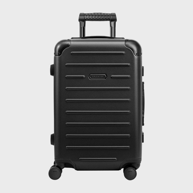 A black suitcase stands upright, featuring a handle and four wheels, against a plain gray background. 