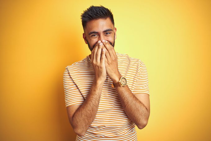 Young Indian Man Wearing a striped tshirt standing in front of a yellow background blushing and covering his face with his hands