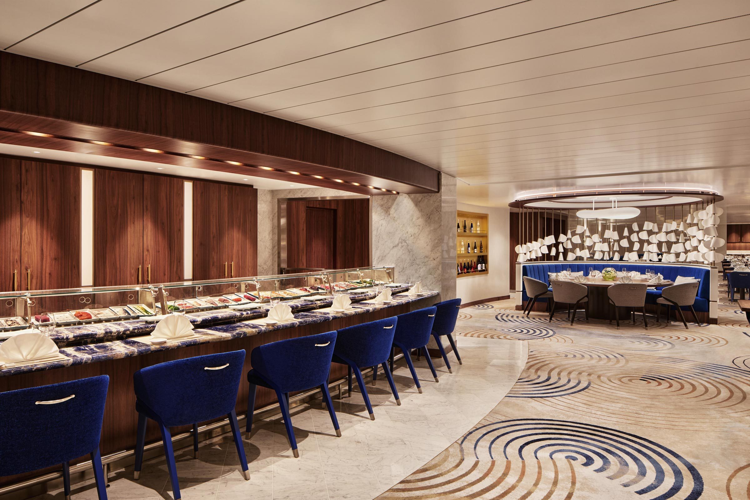 A sushi bar hosts neatly arranged sushi servings, surrounded by blue chairs, within an elegant, modern restaurant with patterned floor and ambient lighting.