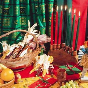 Candles burn on a wooden kinara. Colorful corn and fruits are arranged on a table. A small doll stands nearby, with decorative fabric background.