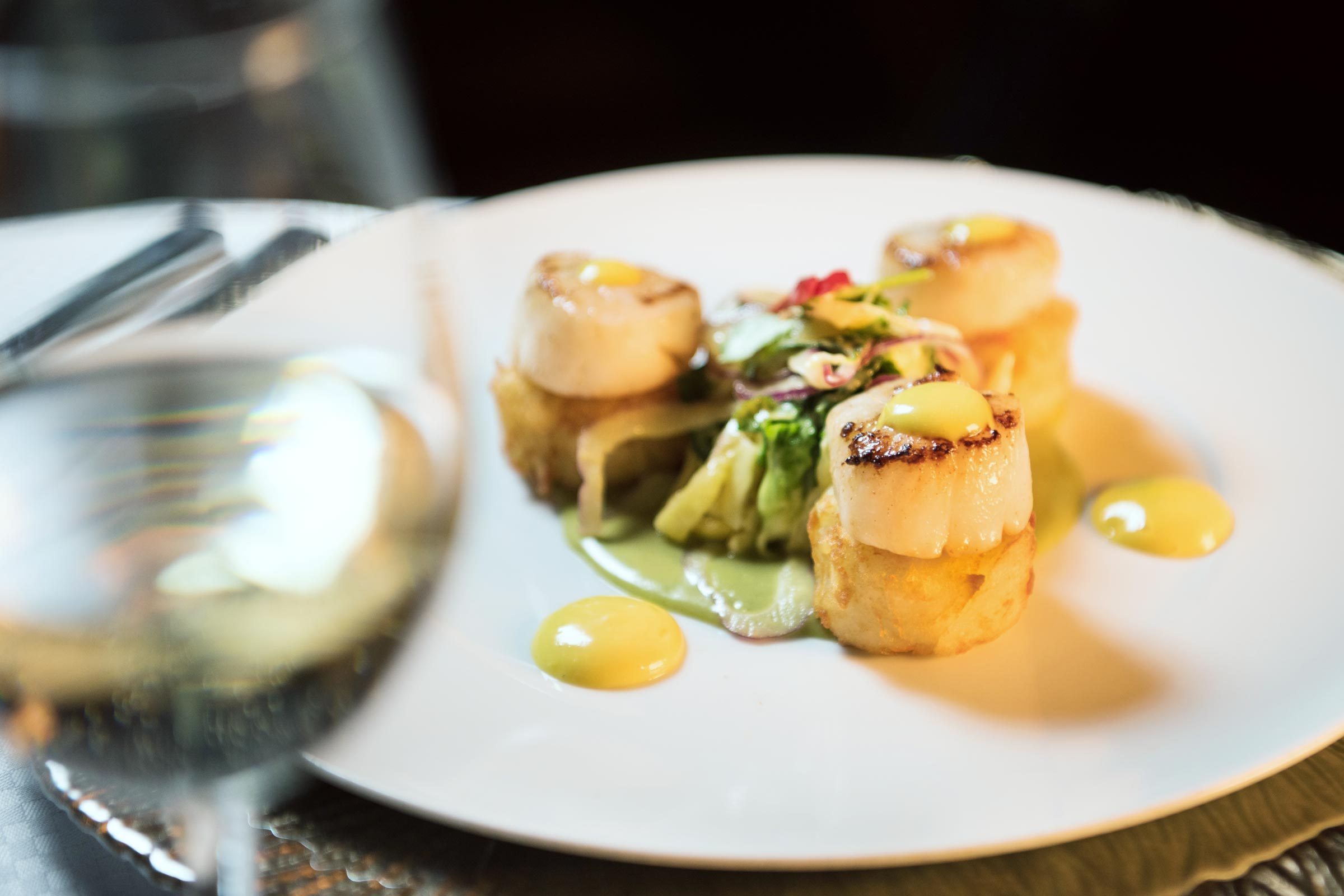 Scallops rest atop potatoes, garnished with sauce and greens on a white plate; a glass of white wine is nearby.