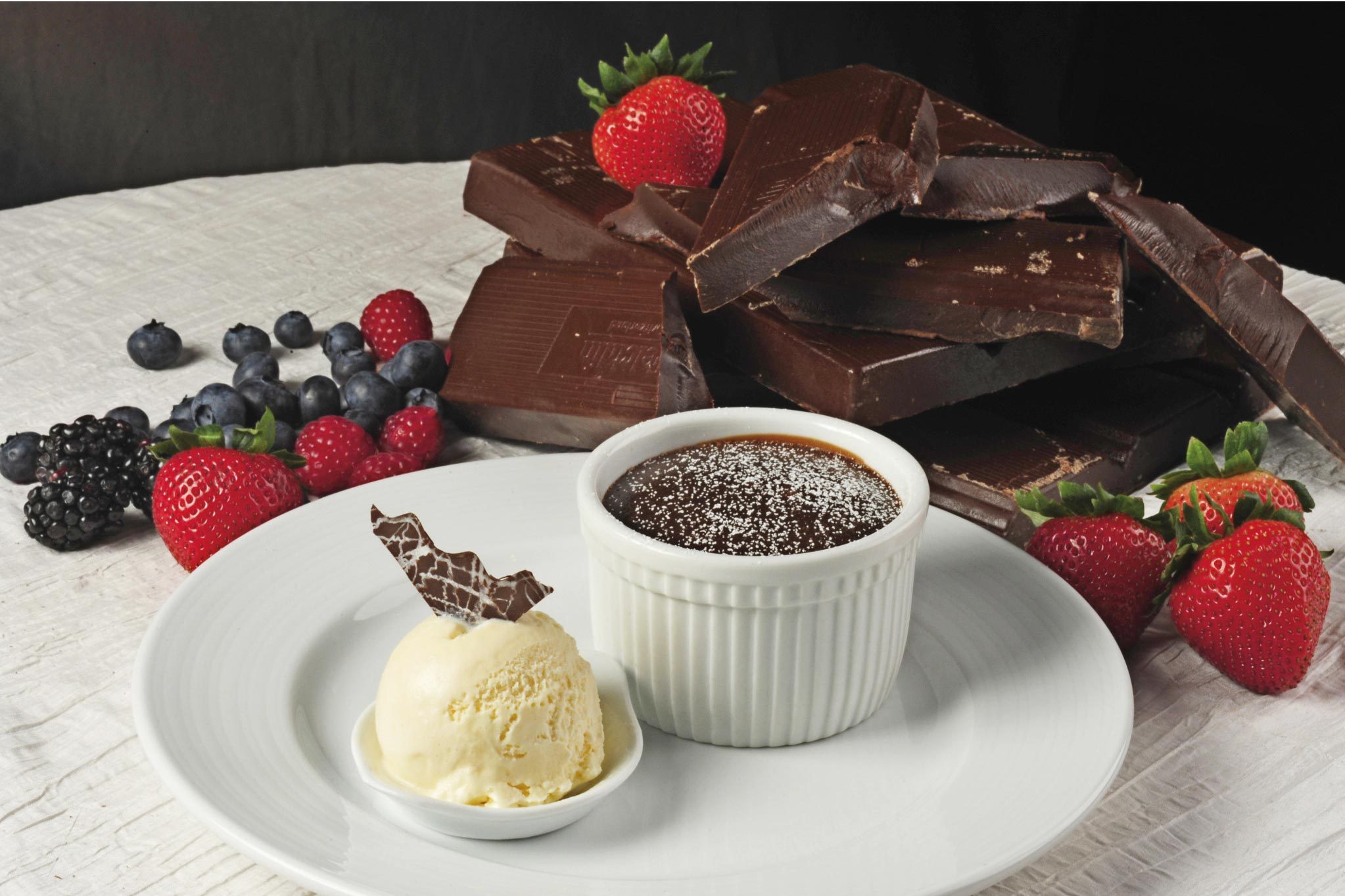 A white ramekin holds chocolate dessert beside vanilla ice cream, surrounded by strawberries, blueberries, blackberries, raspberries, and chocolate blocks on a white tablecloth.