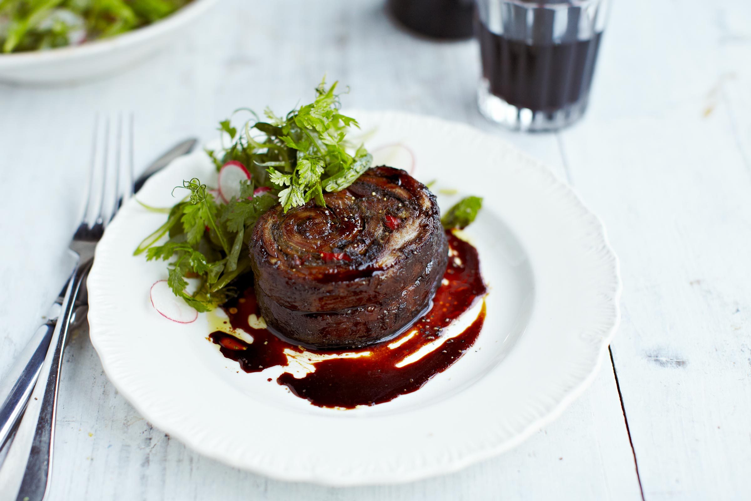 A round, cooked meat with herbs sits on a white plate, drizzled with dark sauce; cutlery and drink nearby, wooden table surface.
