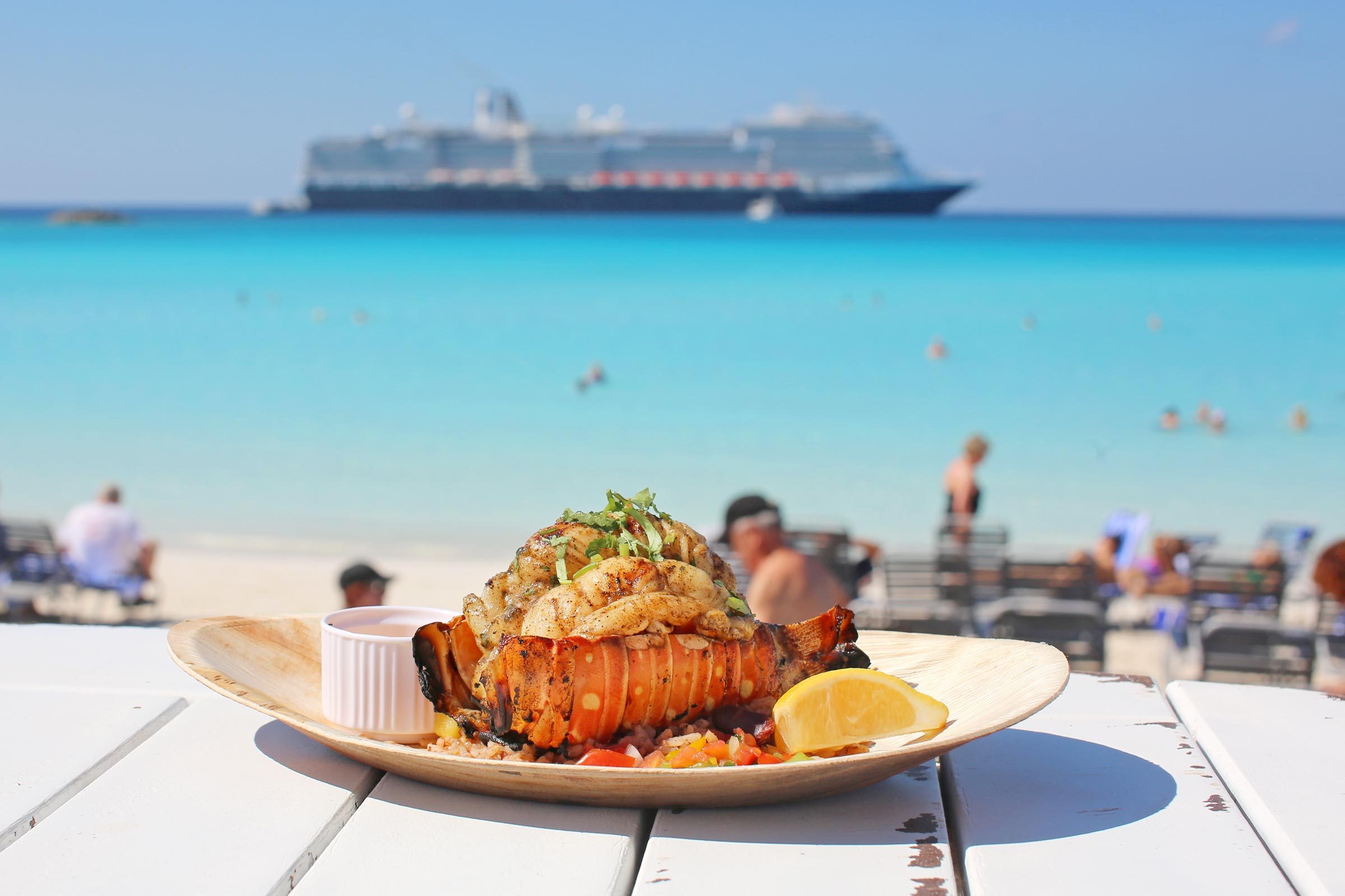 Grilled lobster rests on a plate on a beachside table, with sunbathers and a cruise ship in the turquoise sea background.