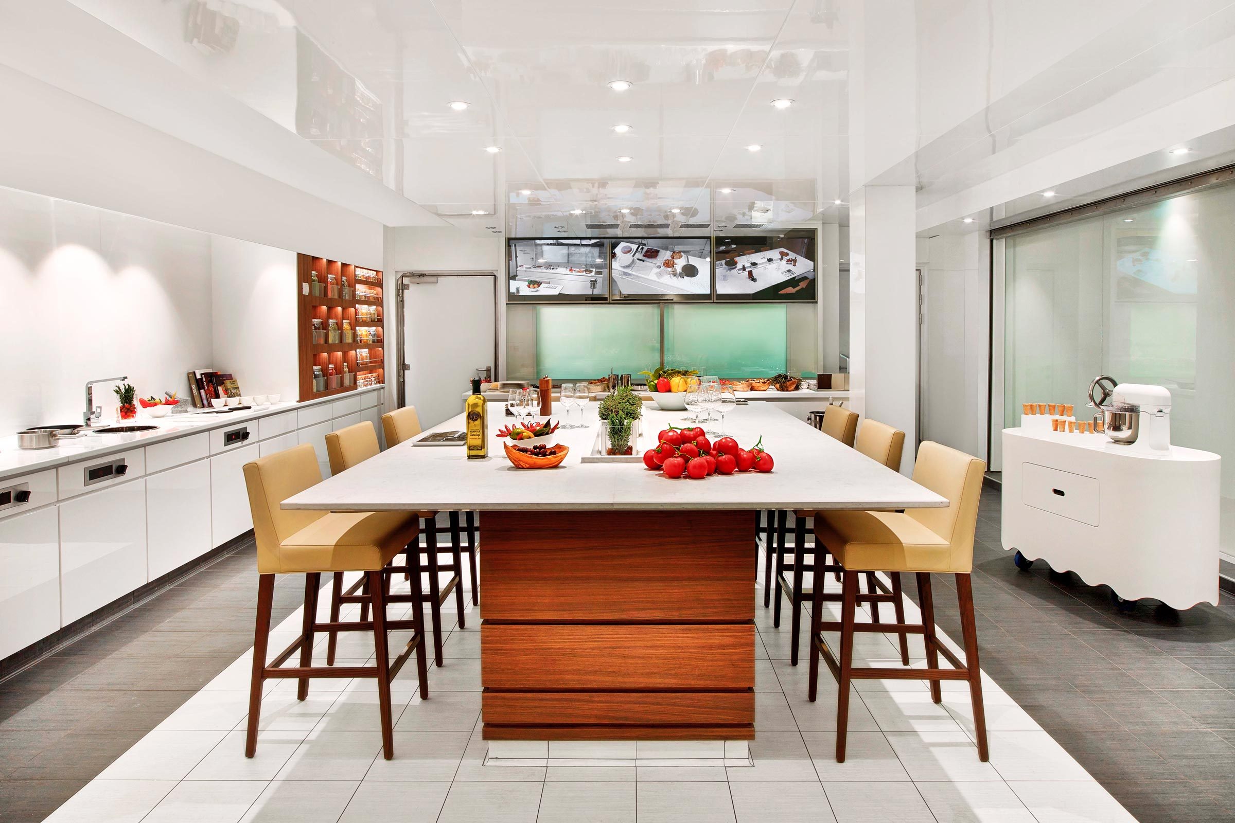 Kitchen island displays fresh vegetables and herbs, surrounded by chairs, within a modern kitchen featuring white cabinets, spice shelves, and overhead screens.