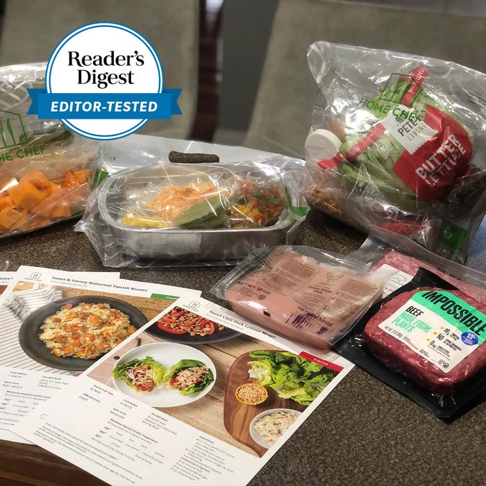 Ingredients displayed on a kitchen countertop accompany recipe cards. Packages include greens, chicken, plant-based meat, and prepared vegetables in a home kitchen setting.