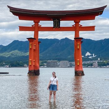 Torii gate stands prominently over water, framing a person, with distant buildings and mountains under a cloudy sky.