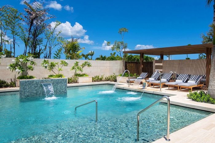 Pool features cascading water, surrounded by lounge chairs under pergola, in a sunny outdoor setting with plants and blue sky.