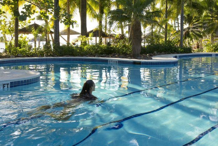 A person swims in a clear blue pool surrounded by lush tropical trees and palm fronds, with sunlight filtering through.