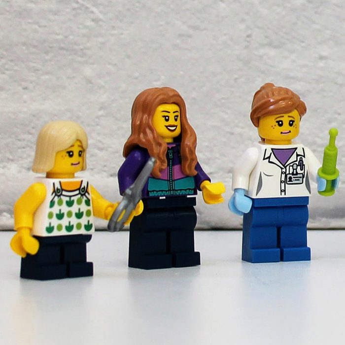 Three LEGO figures stand; one blonde holding scissors, one with long brown hair, and one in a lab coat holding a green syringe.