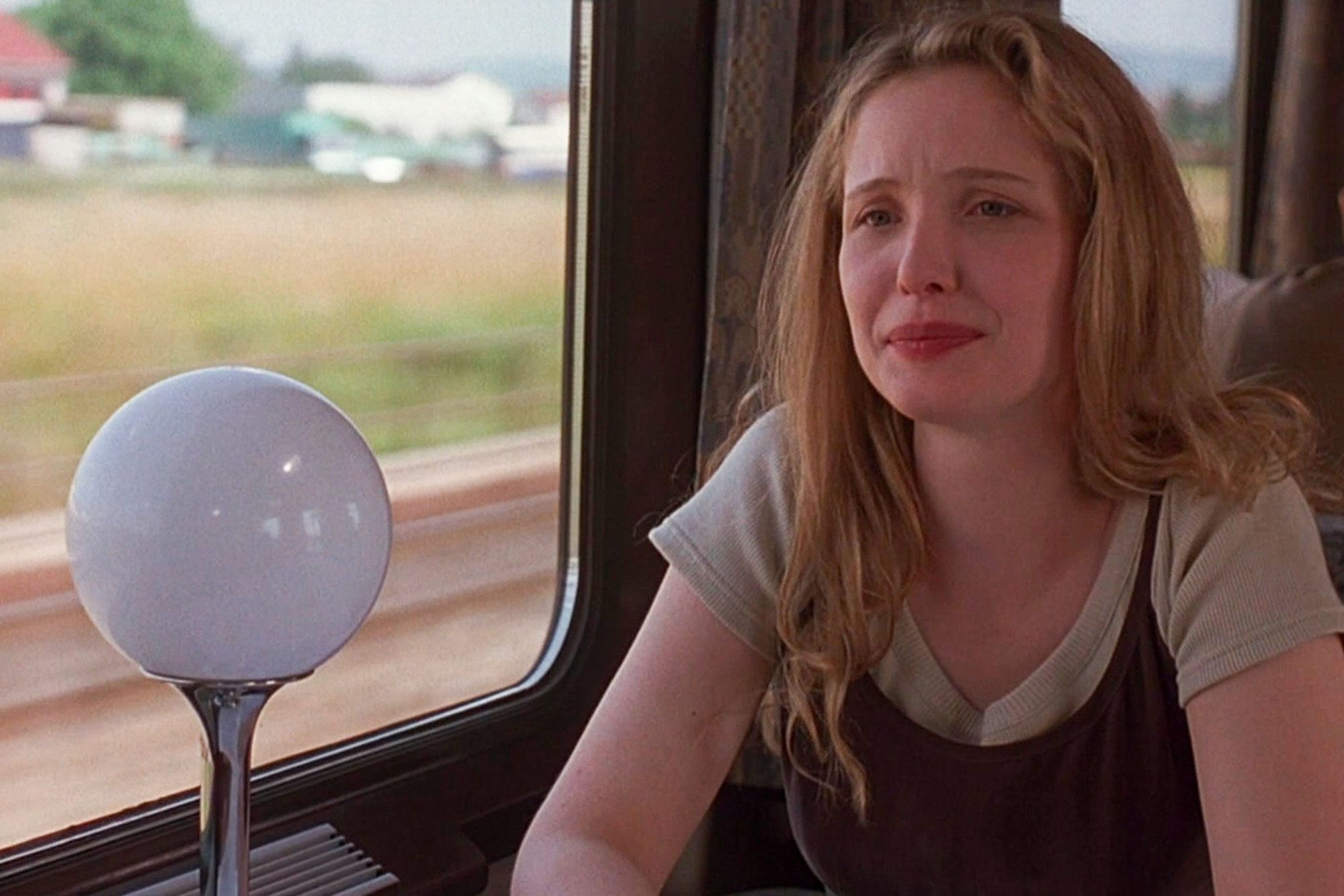A woman sits and gazes out a train window, with a round lamp beside her and fields in the background.