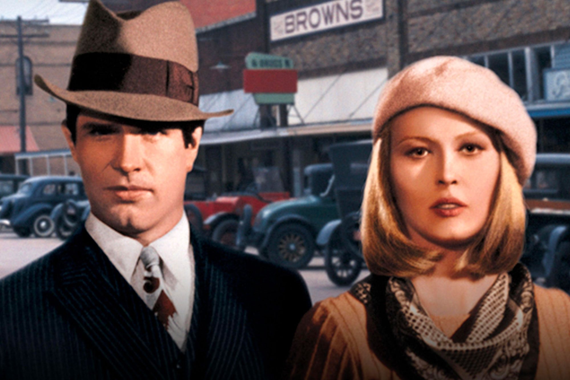 Two people stand, one in a suit and hat, and the other in a beret, in front of vintage cars and a sign reading "BROWNS."
