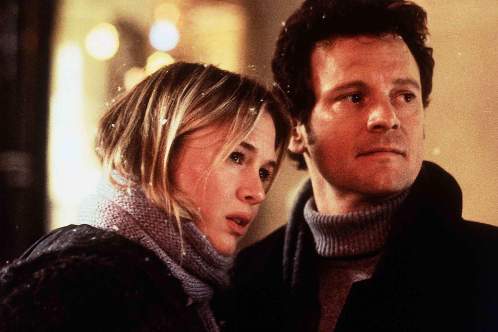 Two people stand closely, looking into the distance, wearing winter clothing in a warmly lit outdoor scene with blurred lights in the background.