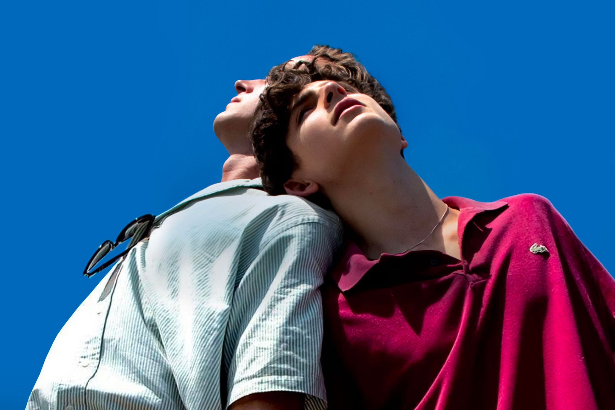 Two individuals lean back-to-back, looking upward, under a clear blue sky. One wears a light shirt with sunglasses; the other, a maroon polo.
