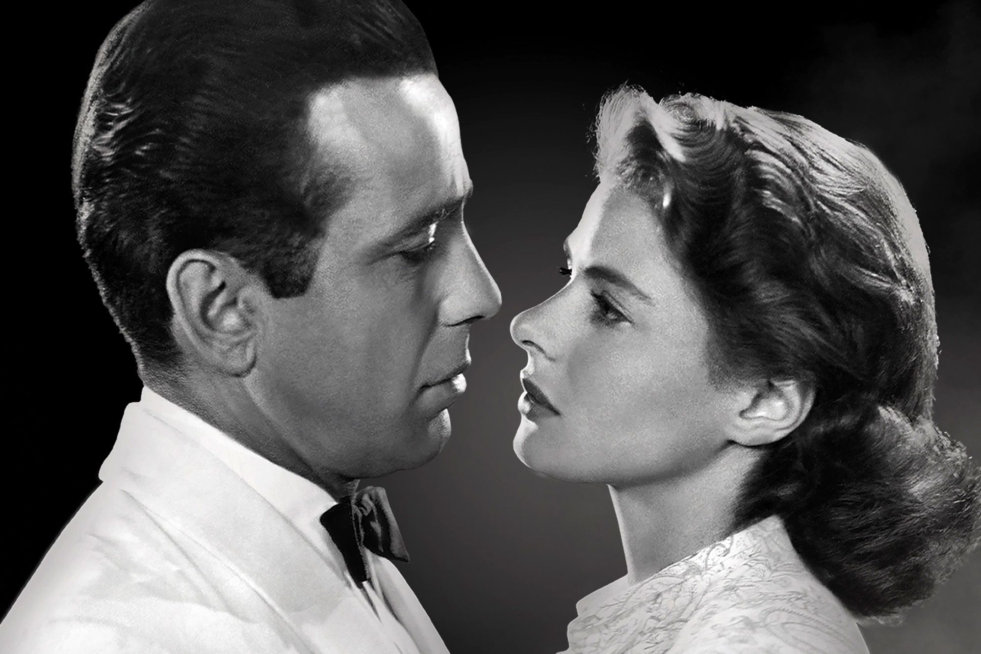 Two people closely face each other, gazing intently. The man wears a suit; the woman wears an elegant blouse. The background is dark and plain.