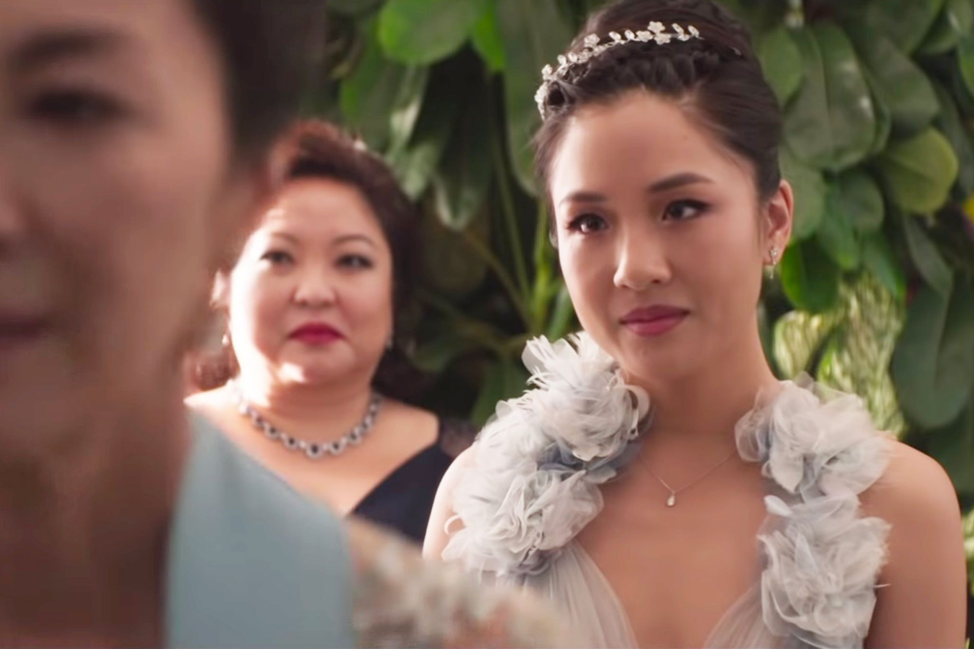 Woman stands serenely, adorned in elegant dress and tiara, in lush, green setting; another woman in background observes, looking intrigued.