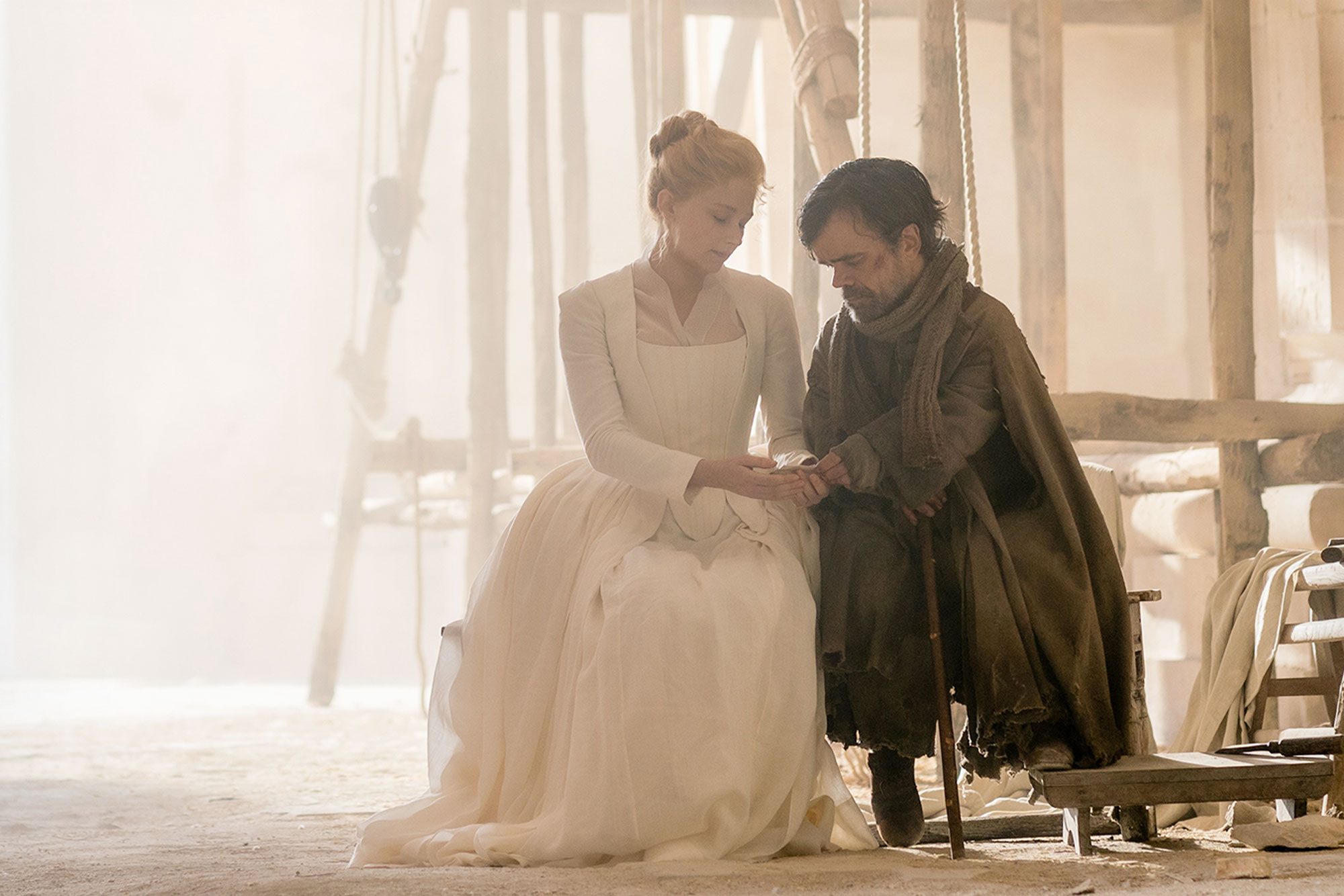 Two people sit closely on a bench, one in white attire, the other in dark clothes, inside a sunlit, rustic room.