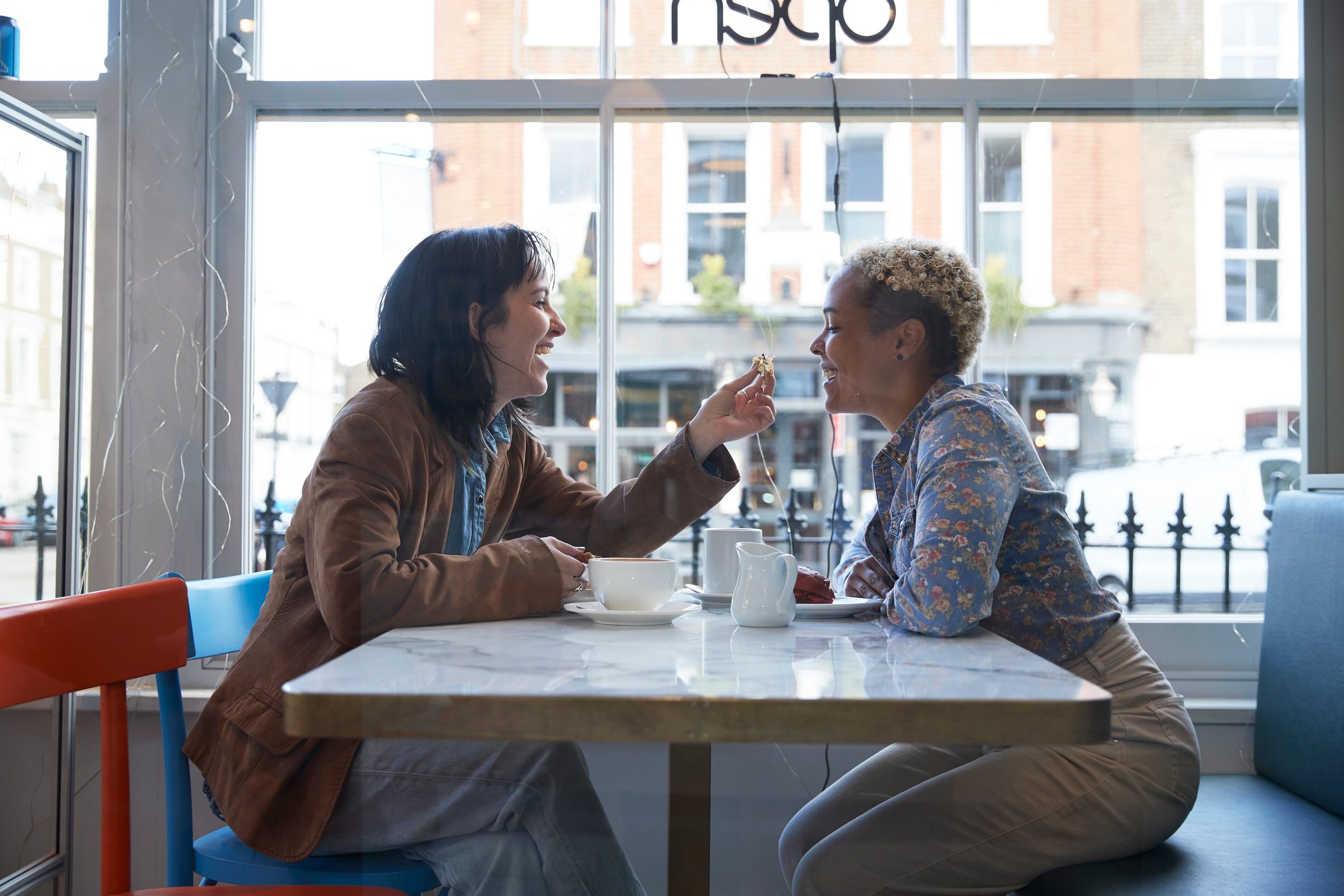 Happy young woman feeding food to girlfriend sitting in cafe