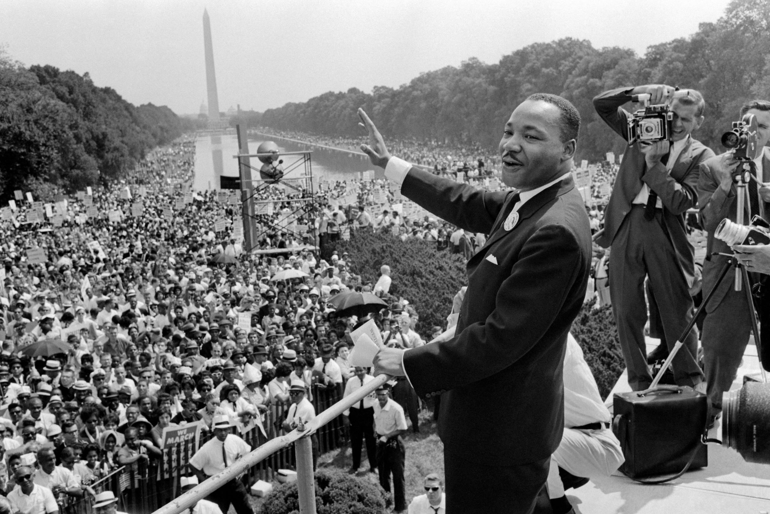 A man waves, addressing a large crowd at a historic civil rights event, with photographers capturing the moment near a reflecting pool and tall monument.