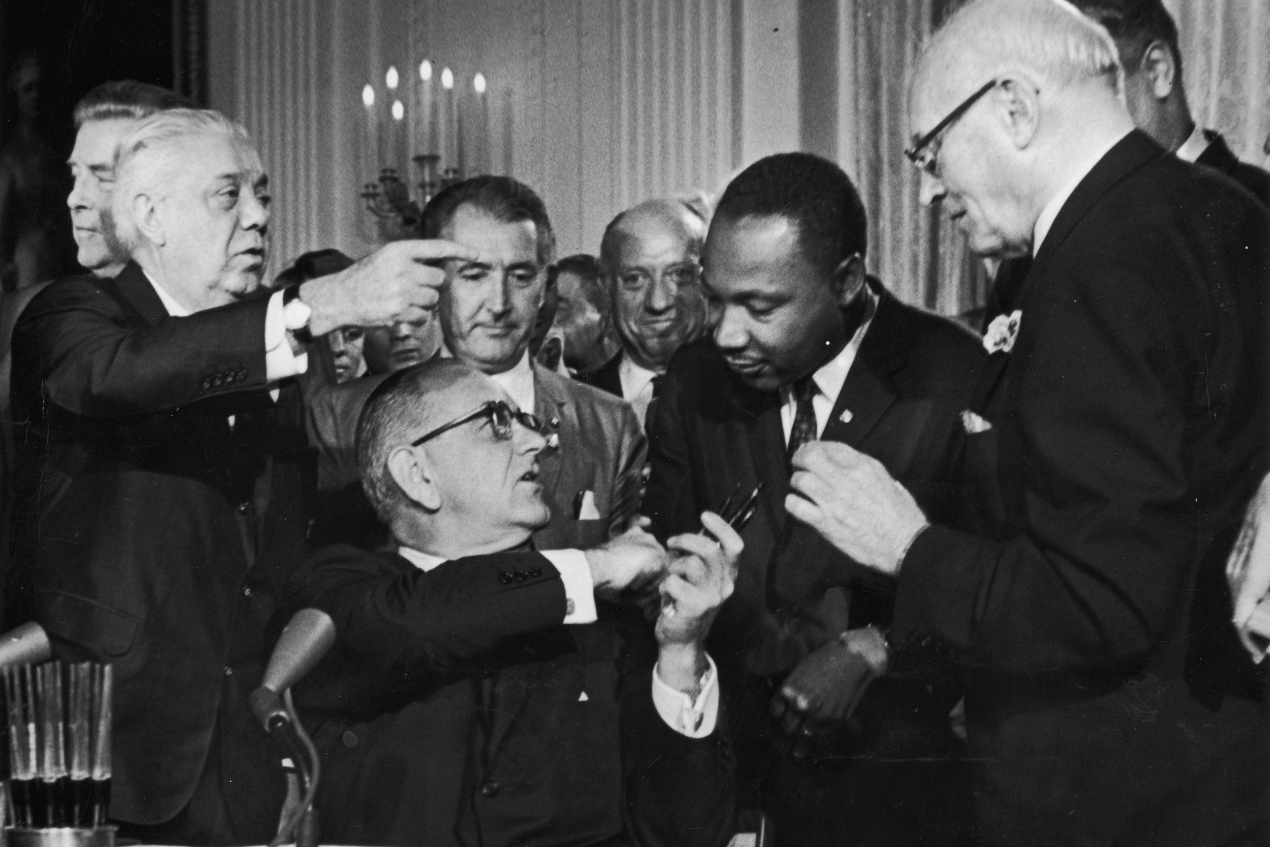 A man sitting hands a pen to another, surrounded by several standing individuals in a formal setting with chandeliers.