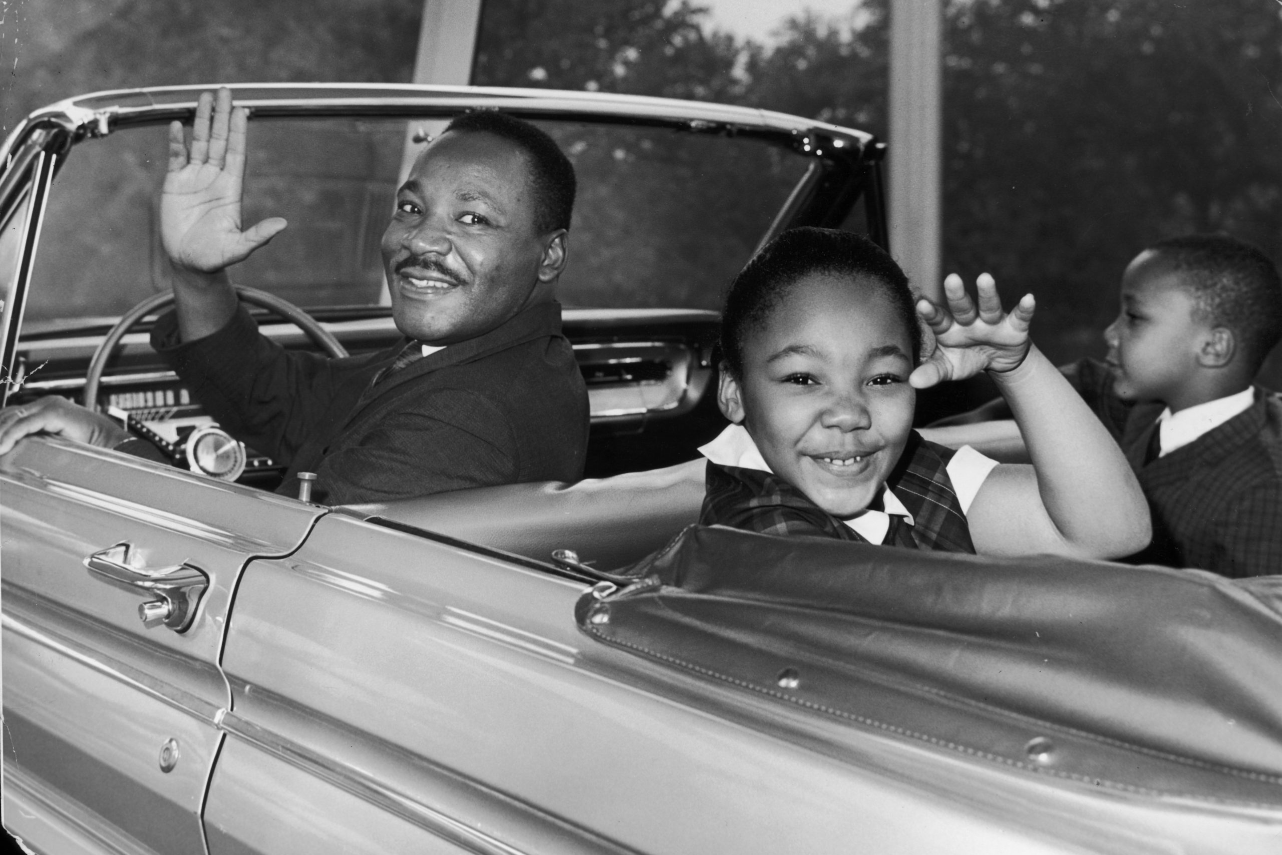 Man driving convertible waves, smiling child in backseat waves, car interior visible.