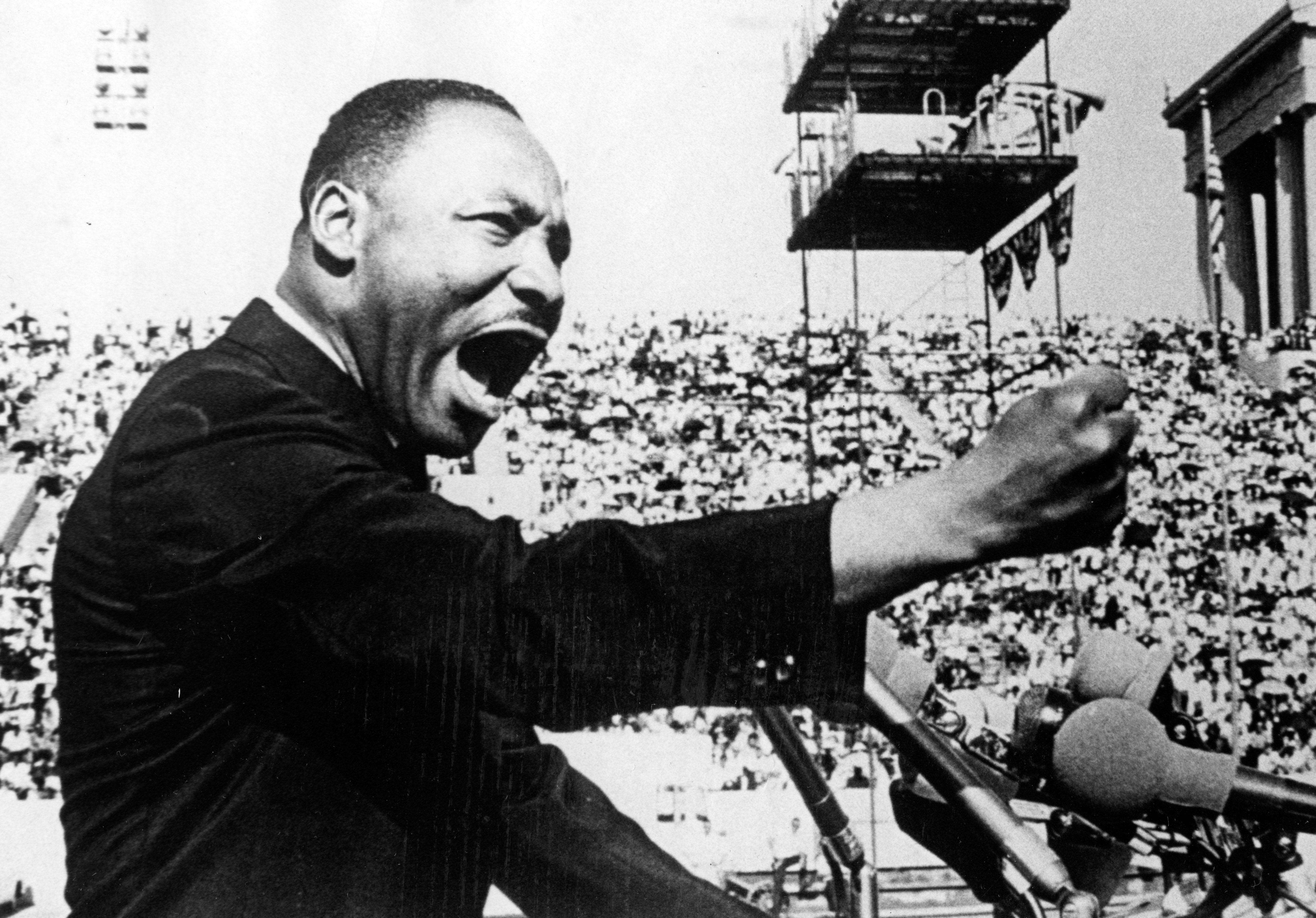 A person passionately speaks into microphones, gesturing with a fist, surrounded by a large, attentive crowd and a prominent building in the background.